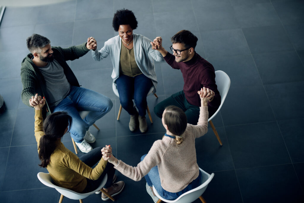 Happy people holding hands while sitting in a circle during group therapy meeting.